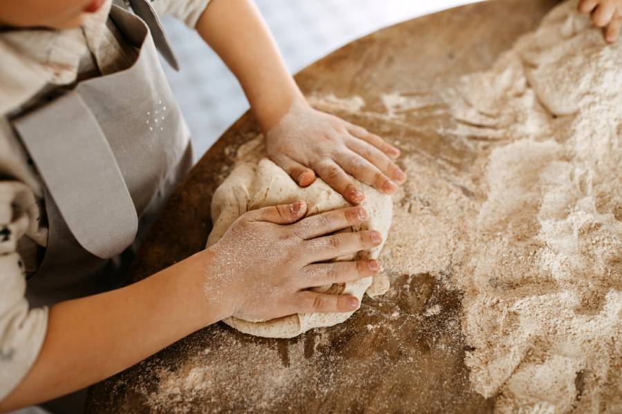 A young child wearing an apron kneading dough on a floured table to make healthy homemade bagels